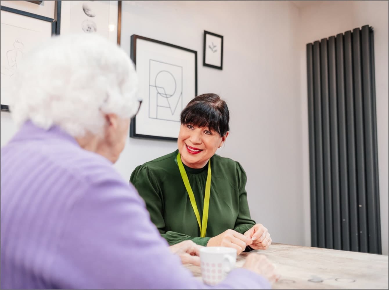 A smiling woman applies makeup to an older woman seated in a bright kitchen with large windows. - Home Instead