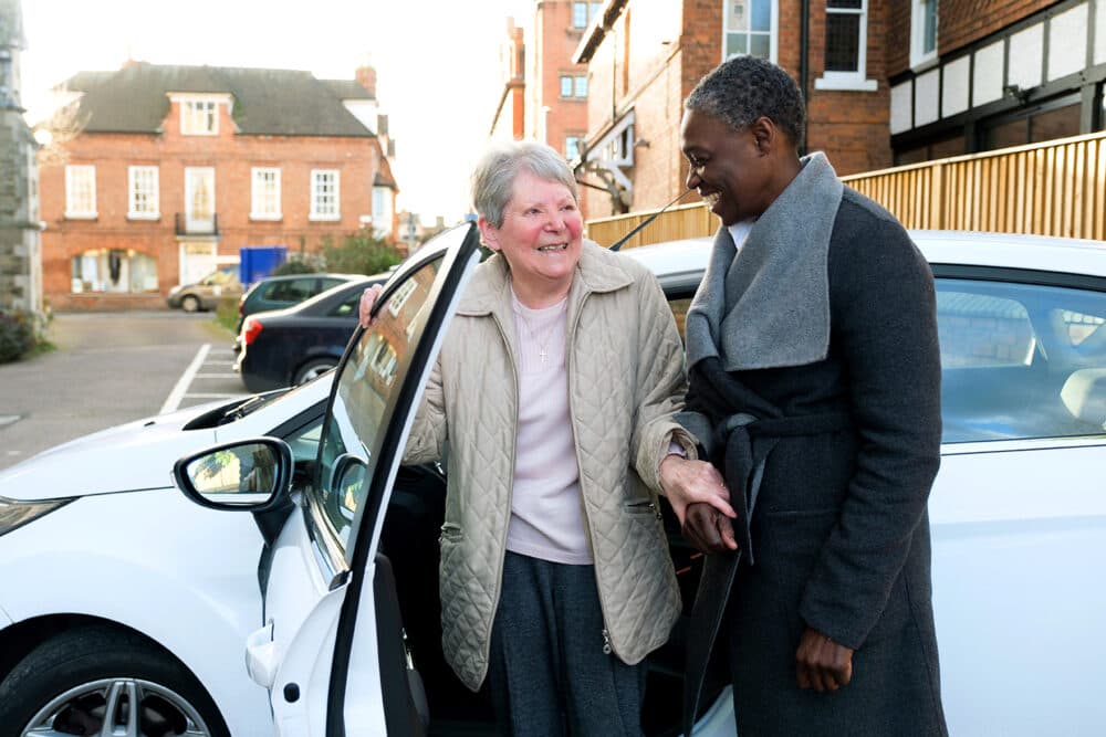 Two women smile as one helps the other get out of a white car in a residential area. - Home Instead