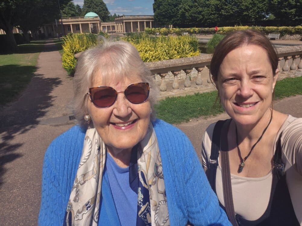 Two women smiling and taking a selfie outside in a sunny garden with pathways and hedges. - Home Instead