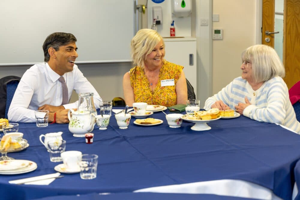Three people sit at a blue-covered table, smiling and talking over tea and pastries in a bright room. - Home Instead