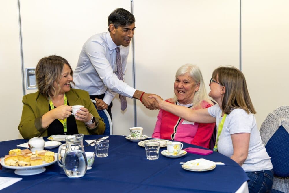 A man in a suit shakes hands with a smiling woman at a table with tea and cakes, among a group of people. - Home Instead