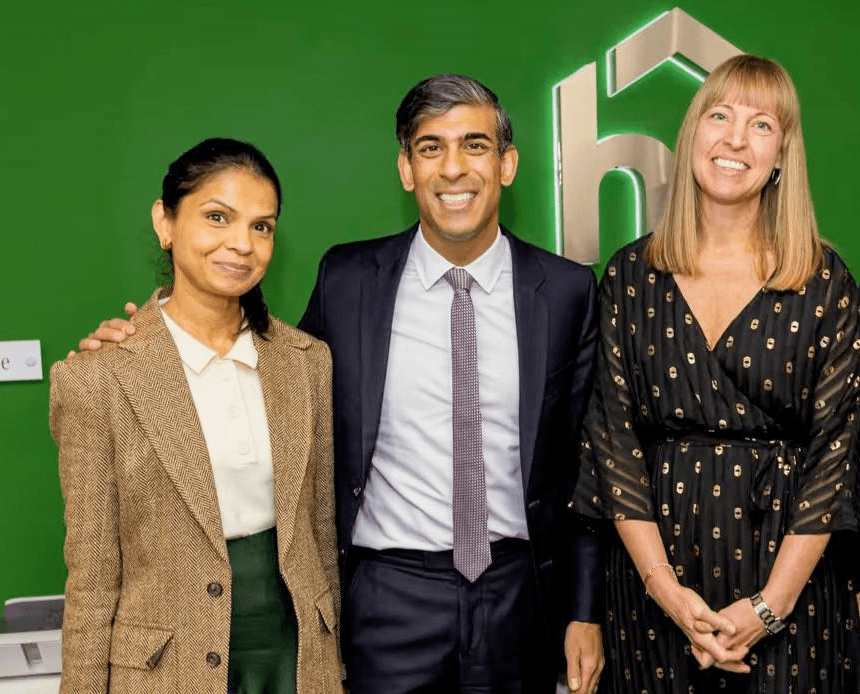 Three people in business attire smiling and posing in front of a green wall with a geometric logo. - Home Instead