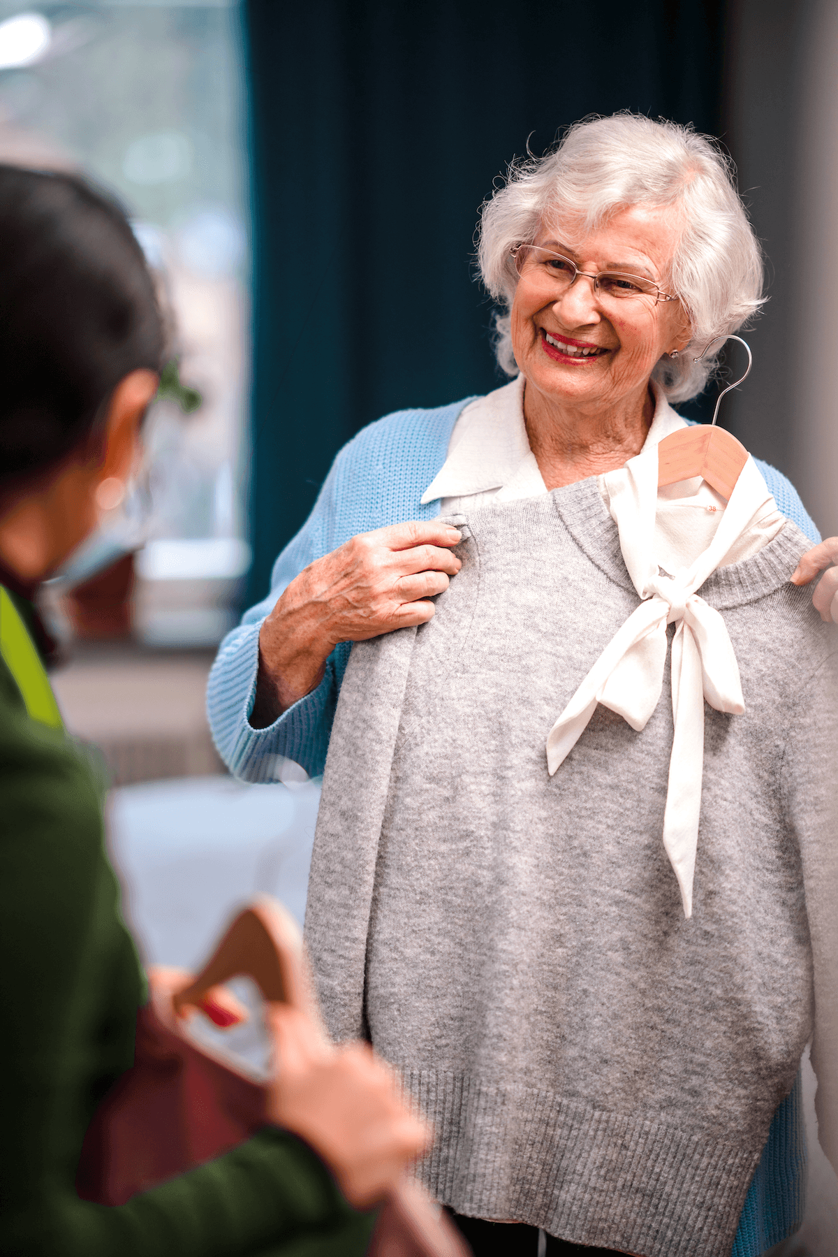 Smiling elderly woman holds a grey jumper on a hanger, showing it to another person indoors. - Home Instead