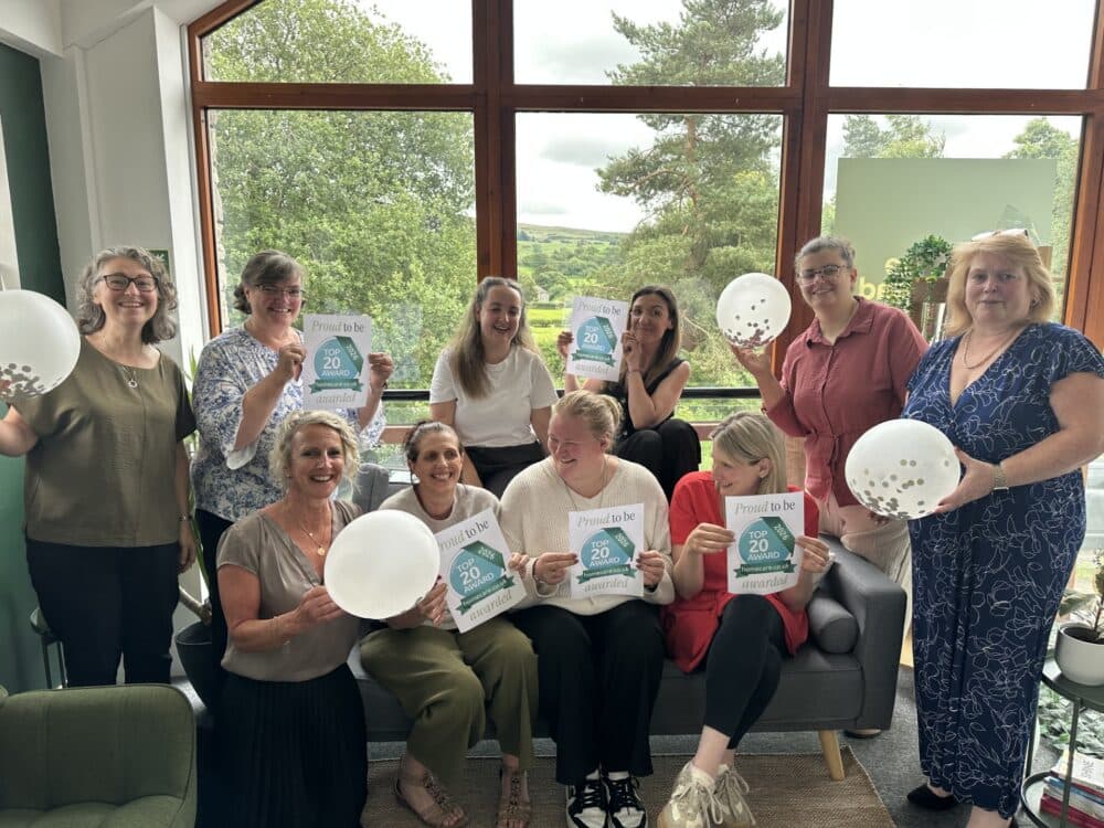 A group of smiling people indoors hold balloons and certificates, celebrating in front of large windows. - Home Instead