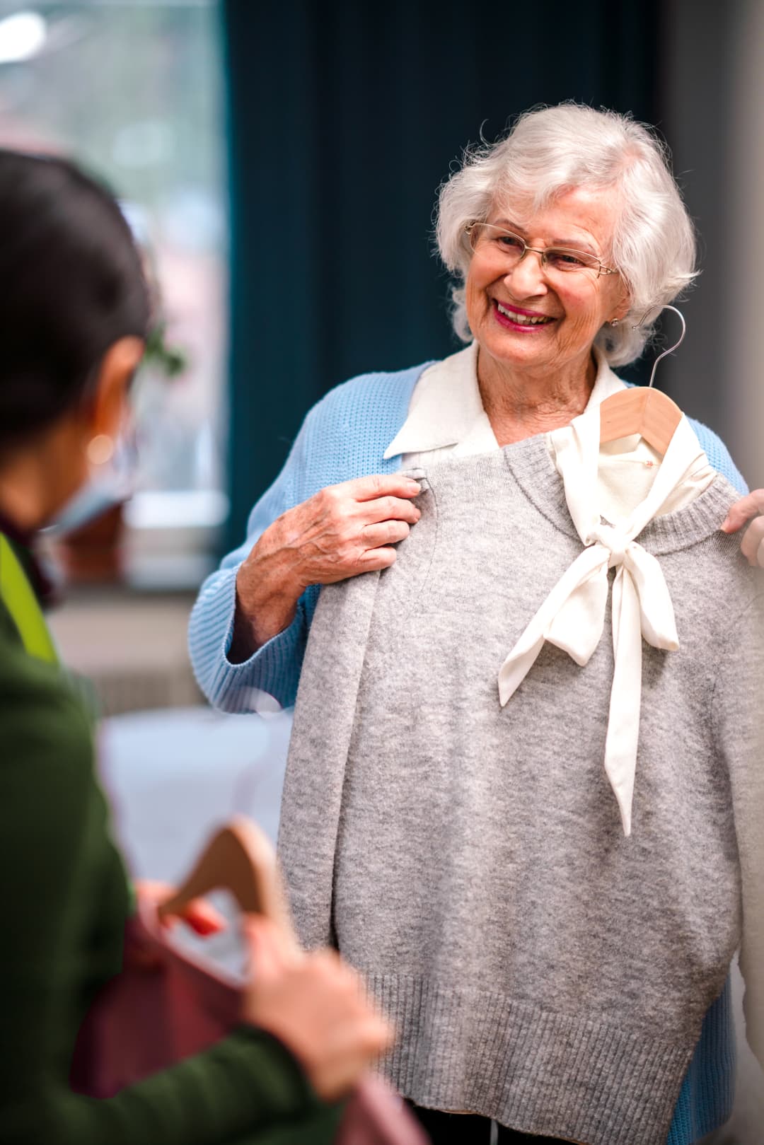 Elderly woman smiling and holding a gray sweater with a white bow while a younger person looks on. - Home Instead