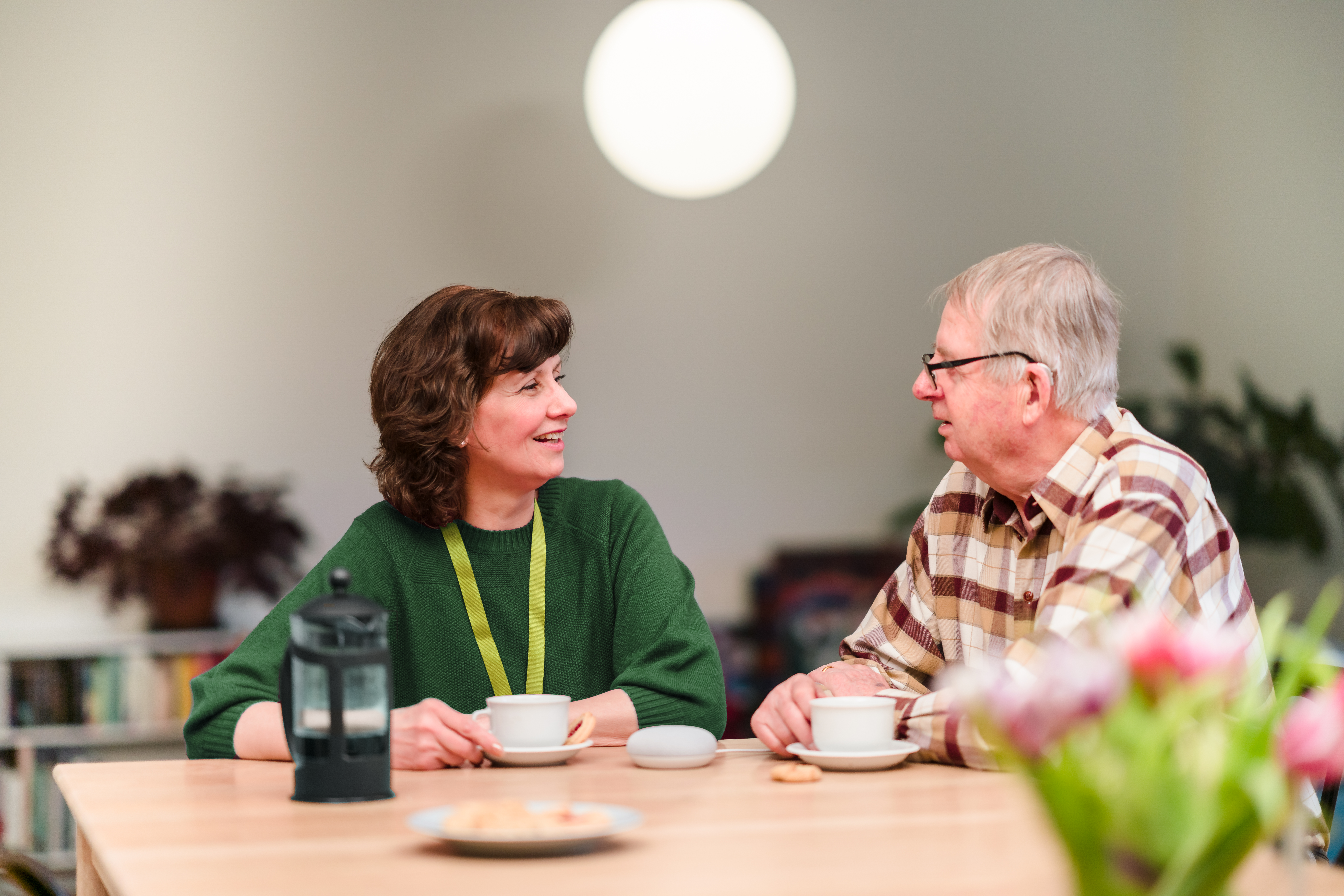An elderly man sitting at a table looking at a Care Professional in a cozy, well-decorated room. - Home Instead