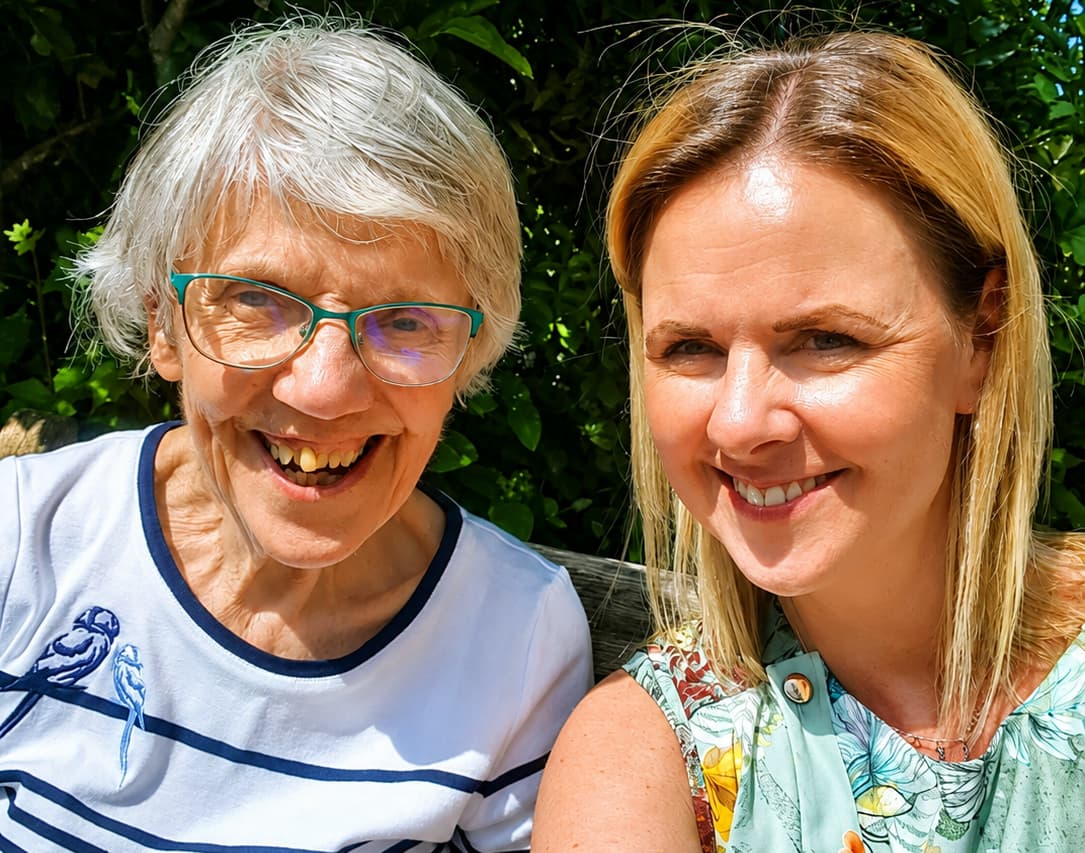 An older woman and a younger woman smiling together outdoors on a sunny day. - Home Instead