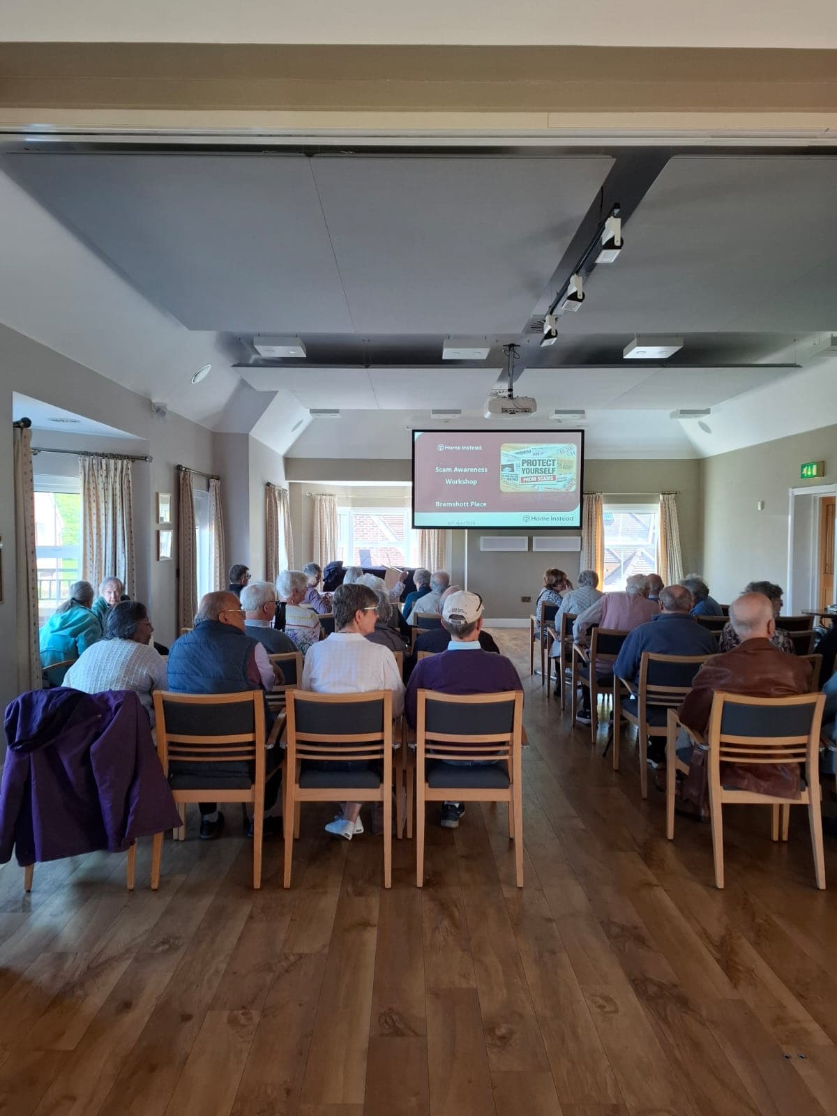 People seated in a community hall, facing a screen displaying a presentation at the front of the room. - Home Instead