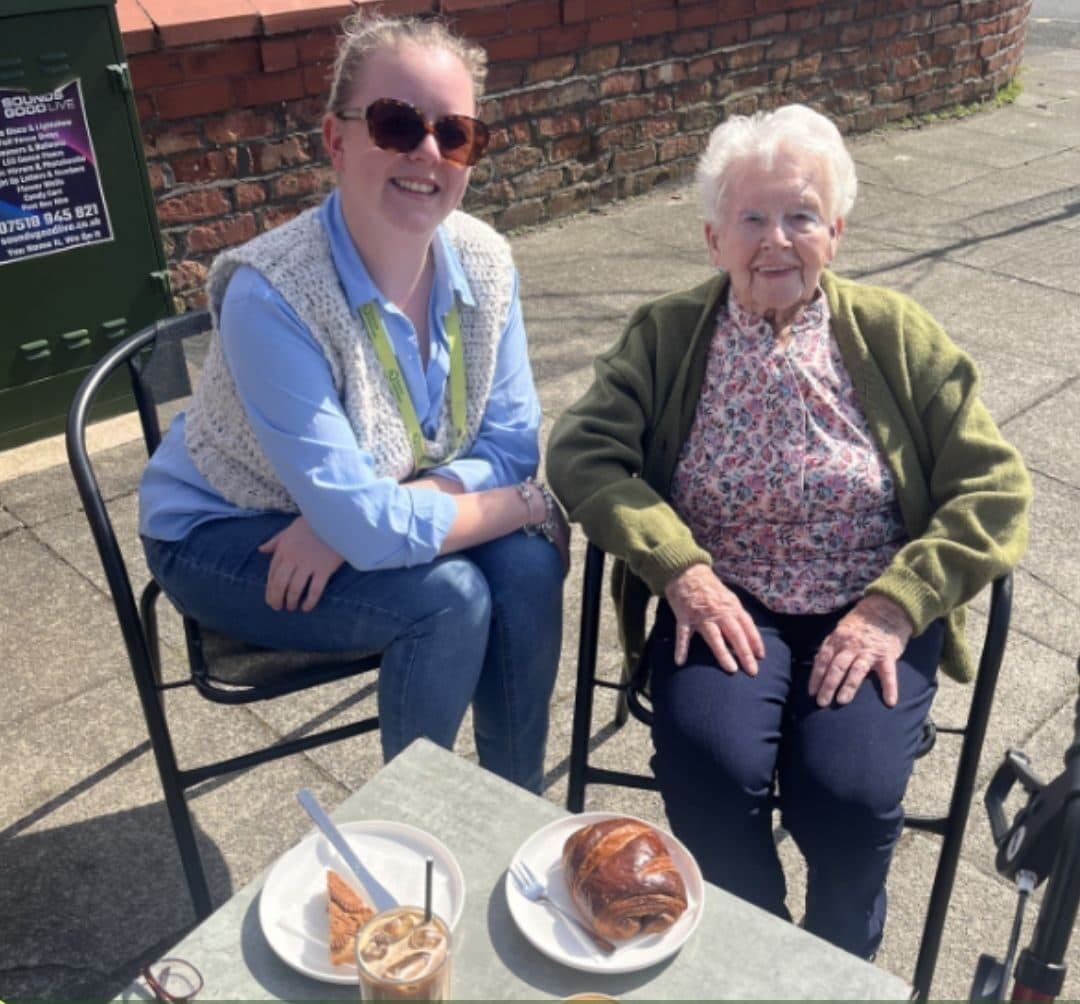 Two women sitting at an outdoor table with pastries and iced coffee, smiling in the sunshine. - Home Instead