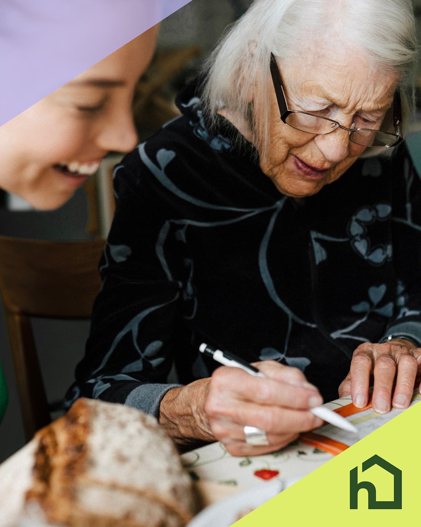 Older woman writing with a pen at a table, smiling person beside her, green house-shaped logo in corner. - Home Instead