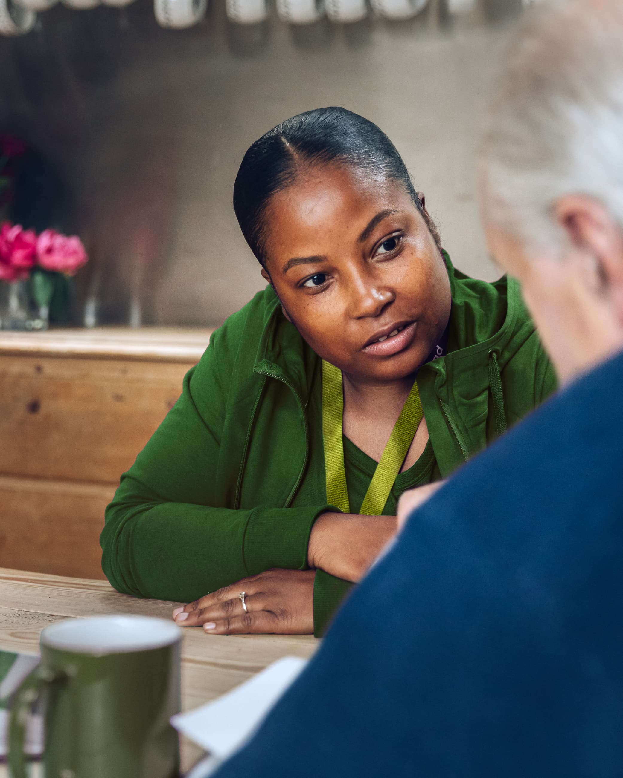 A woman in green listens attentively to an older man across a table in a cosy setting with flowers in the background. - Home Instead