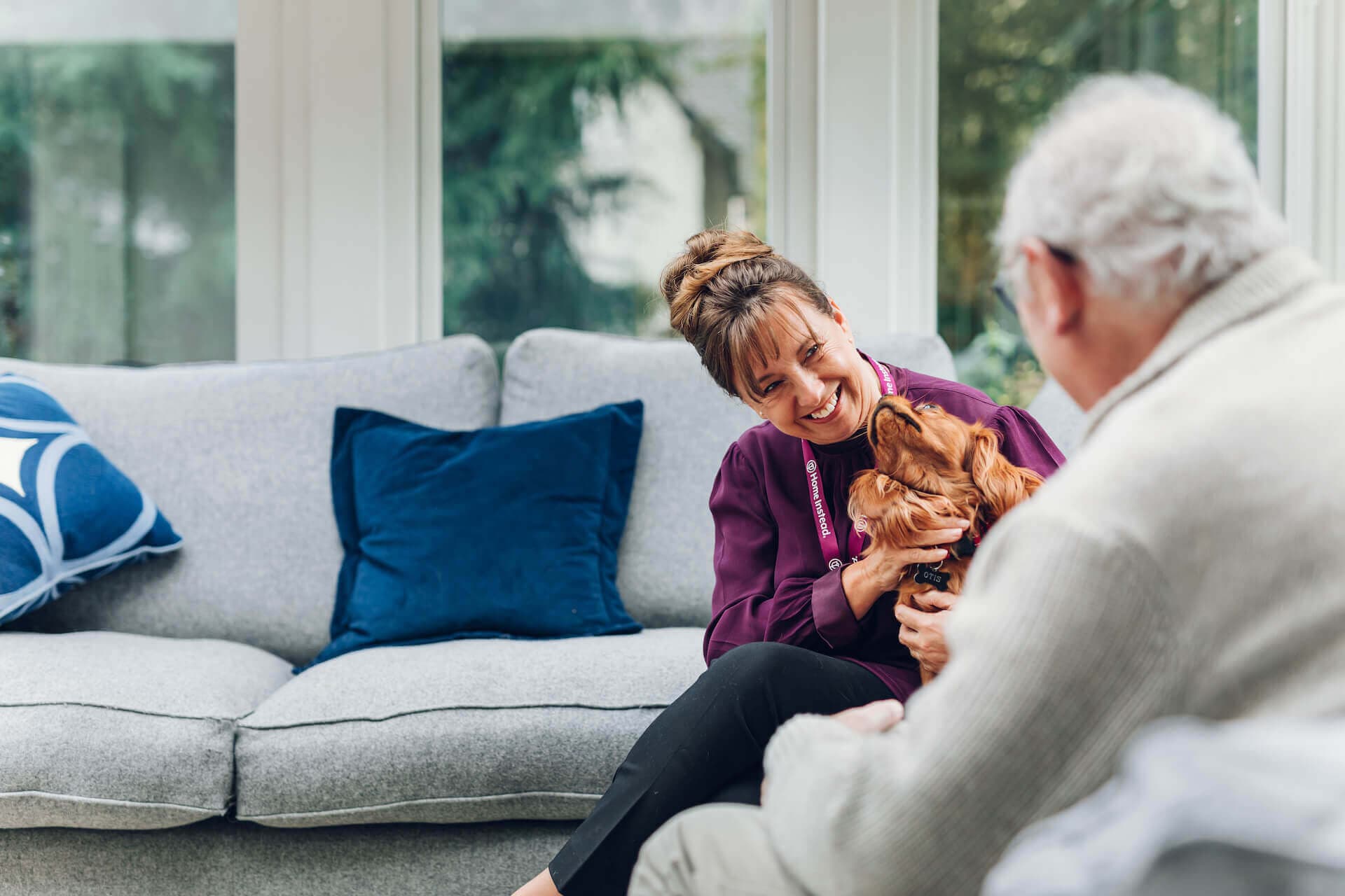 A woman smiles while holding a small dog, sitting on a couch, facing an older man in a cozy living room. - Home Instead