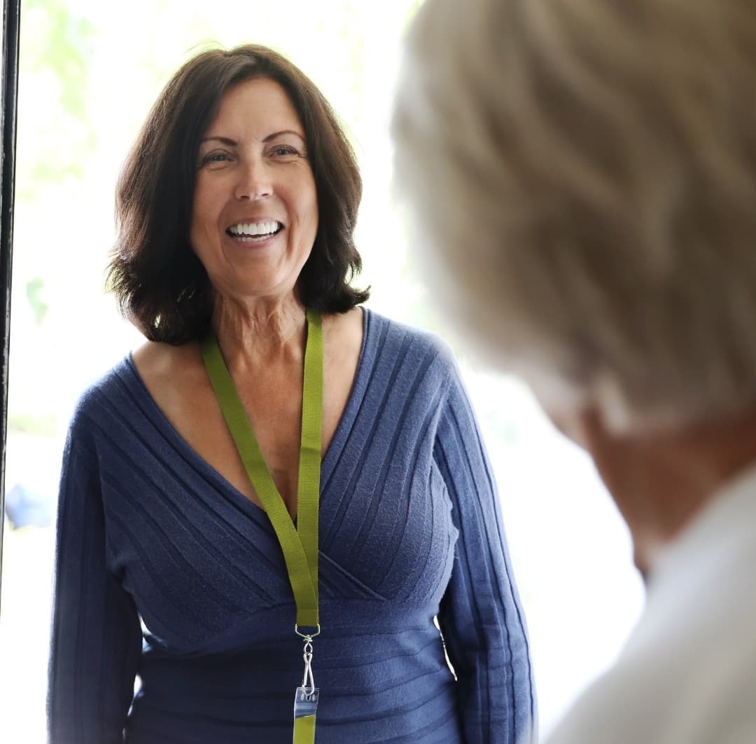 Smiling woman with a lanyard greets another person at a door. - Home Instead