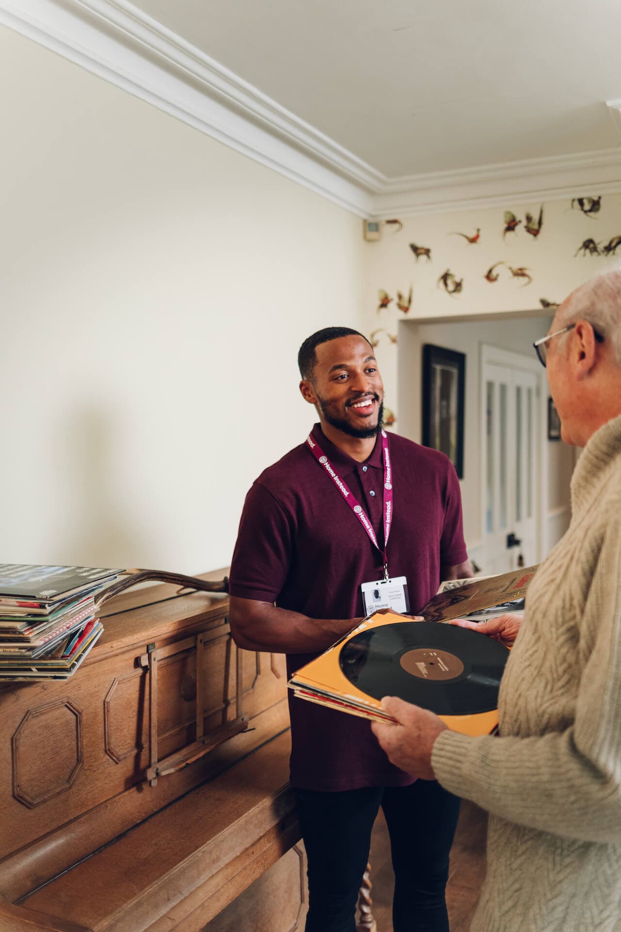 Two people exchange vinyl records indoors near a piano, with one wearing a name badge and smiling. - Home Instead