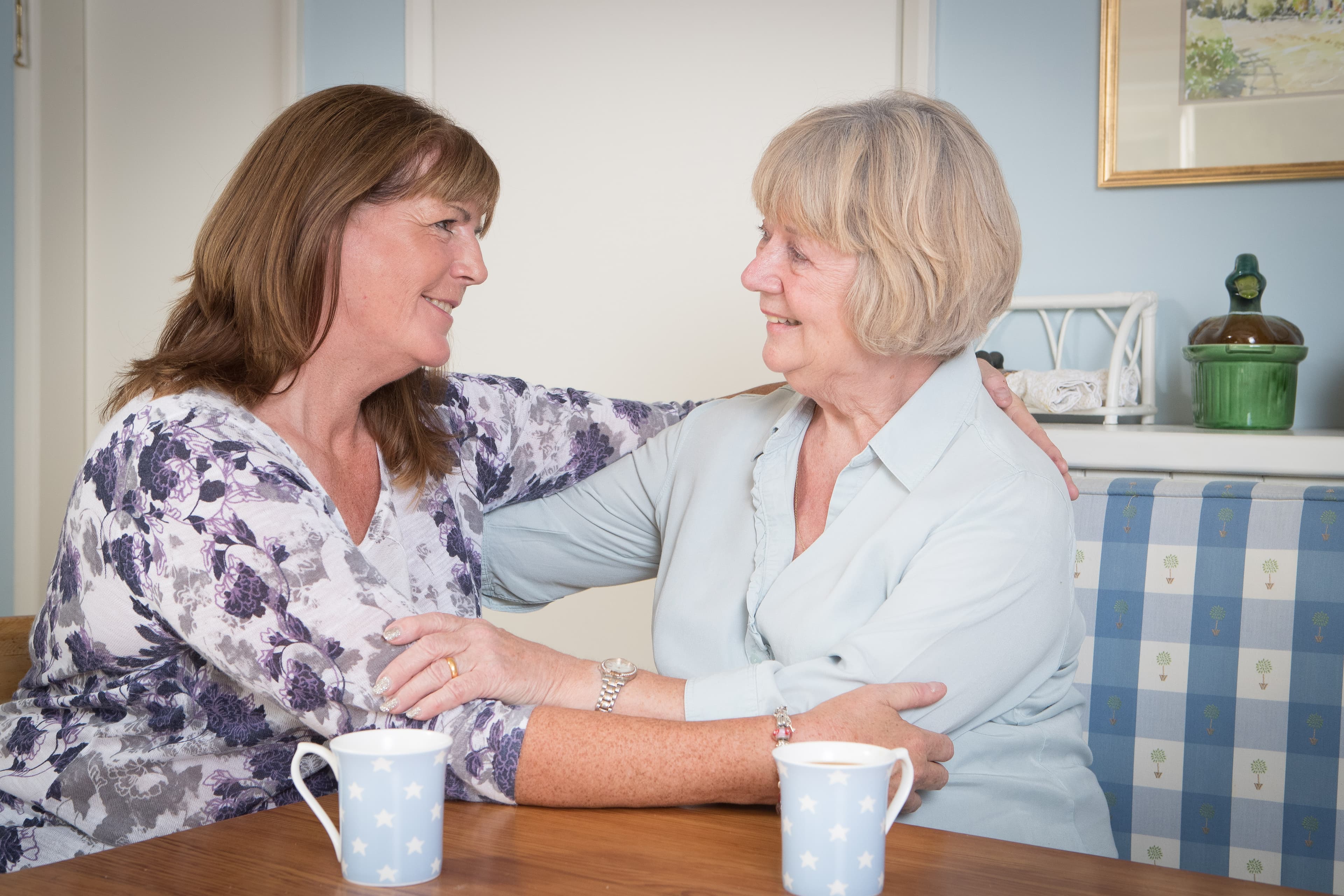Two women smiling at each other, sitting at a table with two mugs, in a light, cozy room. - Home Instead