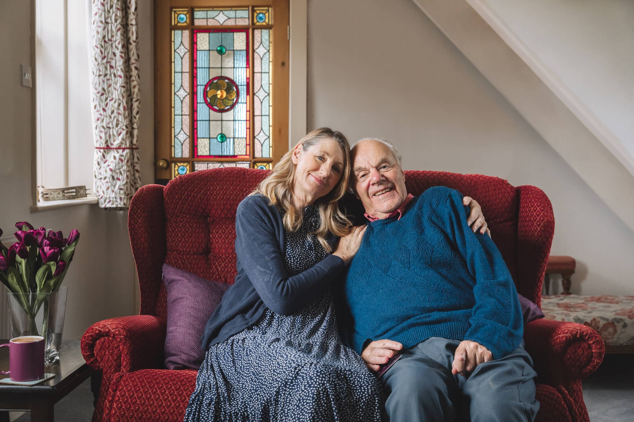 An elderly couple sits on a red couch, smiling warmly. Stained glass window and flowers are in the background. - Home Instead