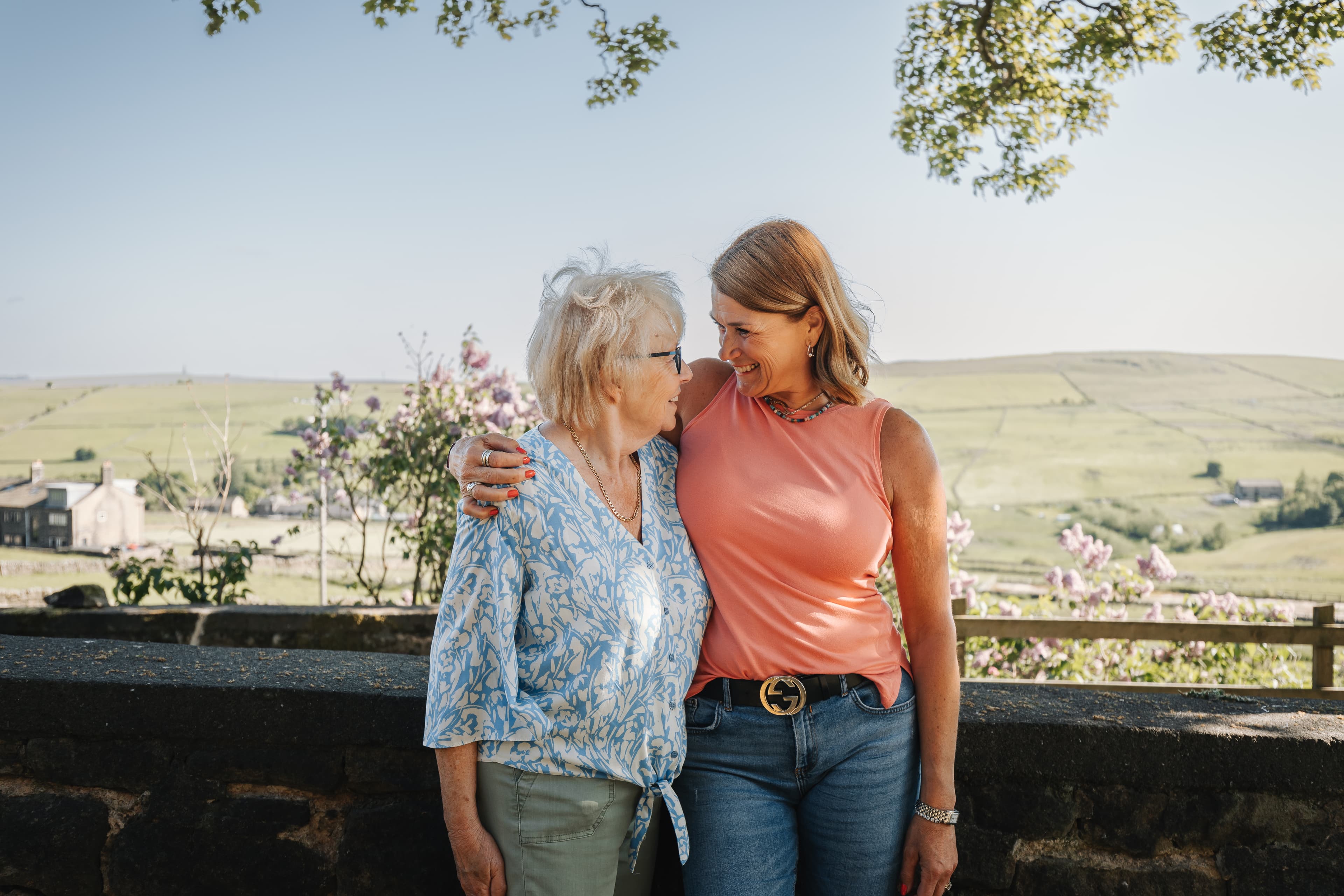 An older woman and a younger woman smiling and hugging outdoors with fields in the background. - Home Instead