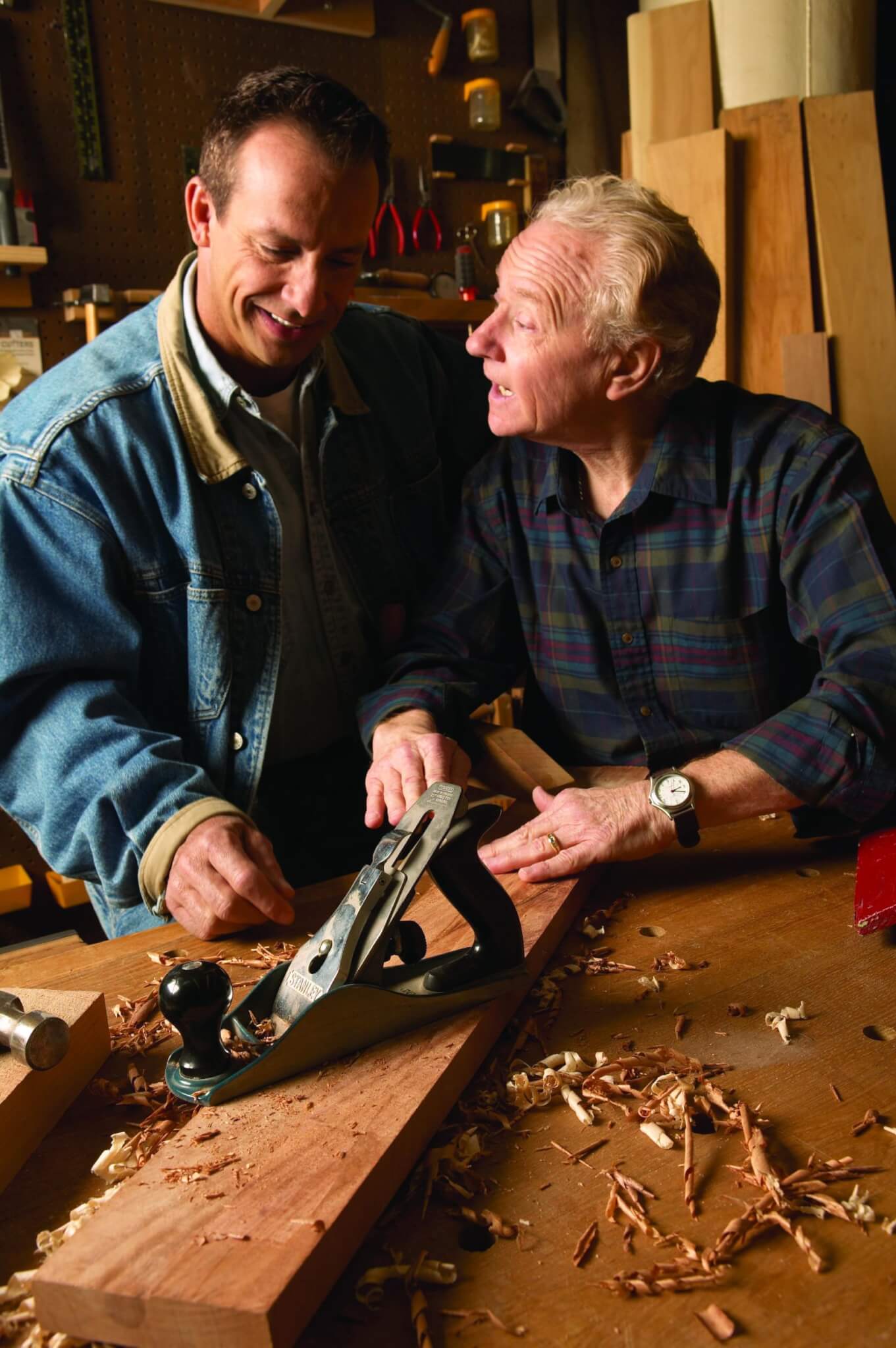 Two men in a workshop, one teaching the other how to use a hand plane on a piece of wood. - Home Instead