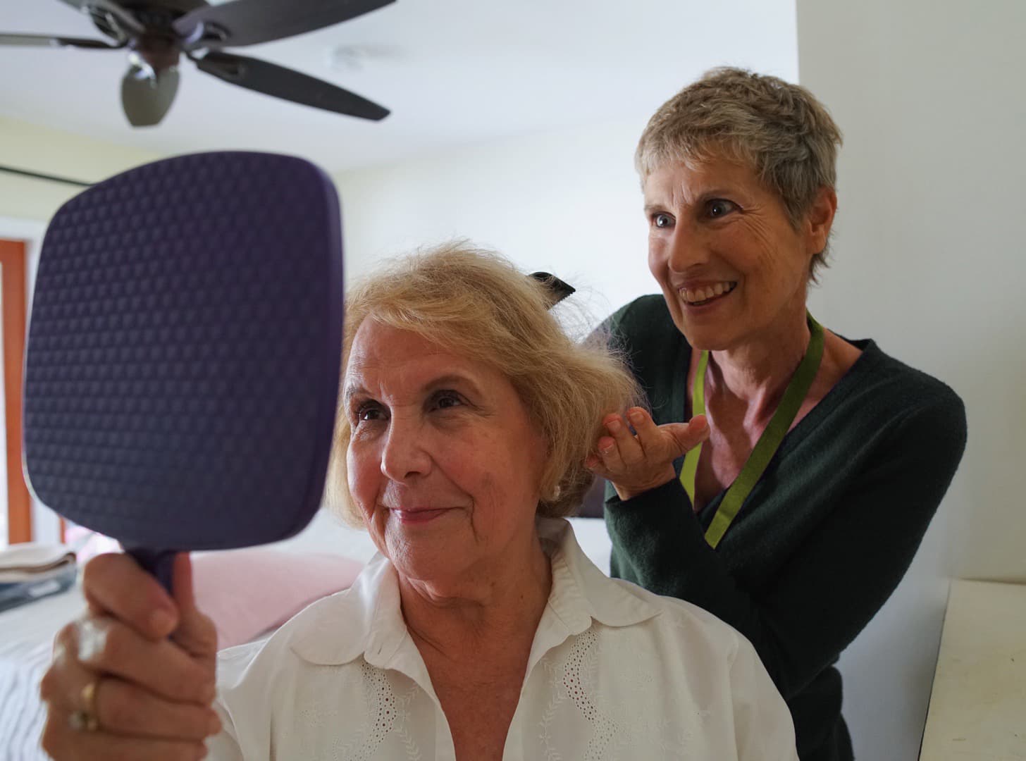 An older woman smiles at a hand mirror while another woman stands behind her, styling her hair. - Home Instead