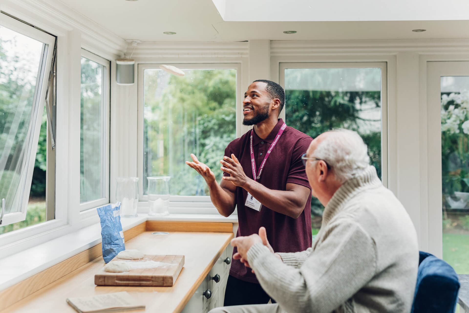 A young man and an elderly man chatting and gesturing in a bright kitchen with large windows. - Home Instead