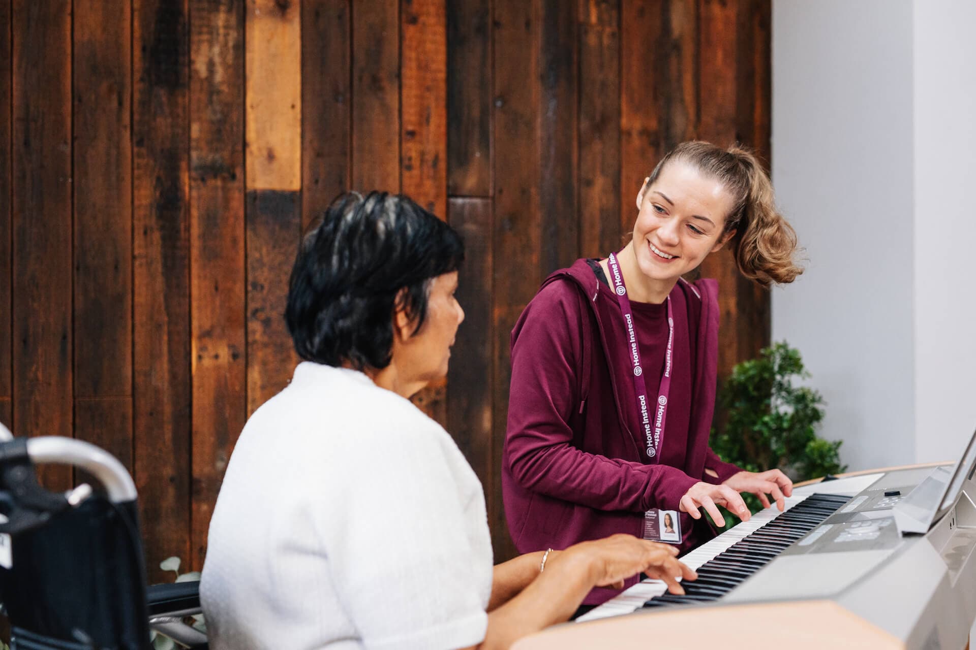 A young woman smiles while playing the keyboard with an elderly woman in a white sweater against a wood-paneled background. - Home Instead