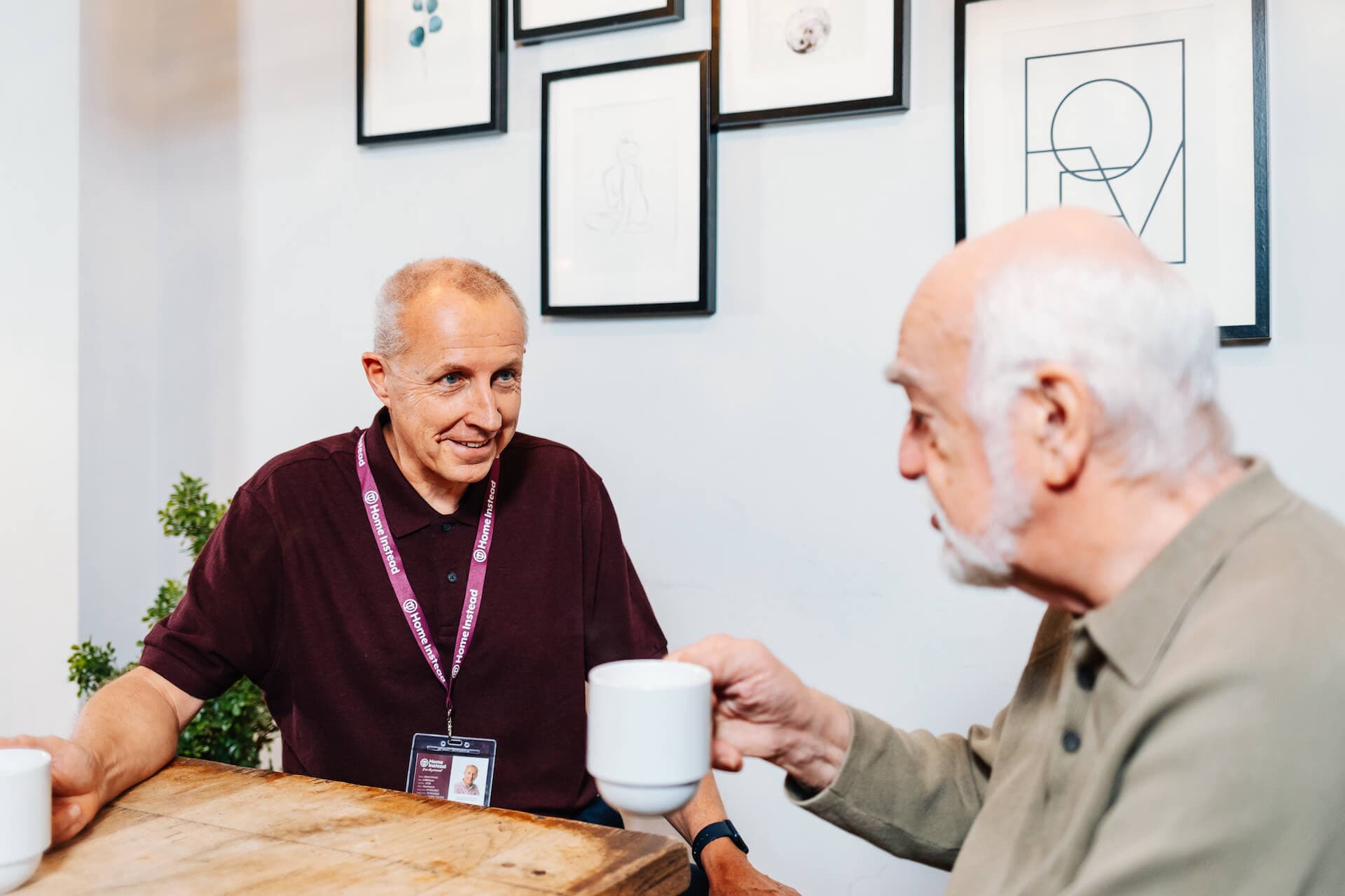 Two senior men sitting at a table with coffee mugs, engaging in conversation with framed art on the wall behind them. - Home Instead
