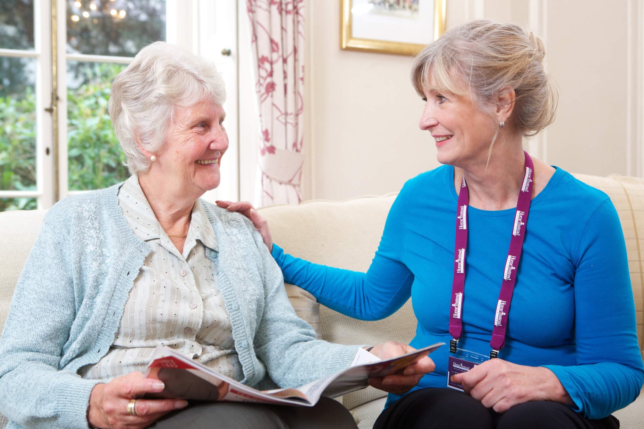 An elderly woman and a caregiver sit on a couch, smiling and talking, with a magazine in the elderly woman’s hands. - Home Instead