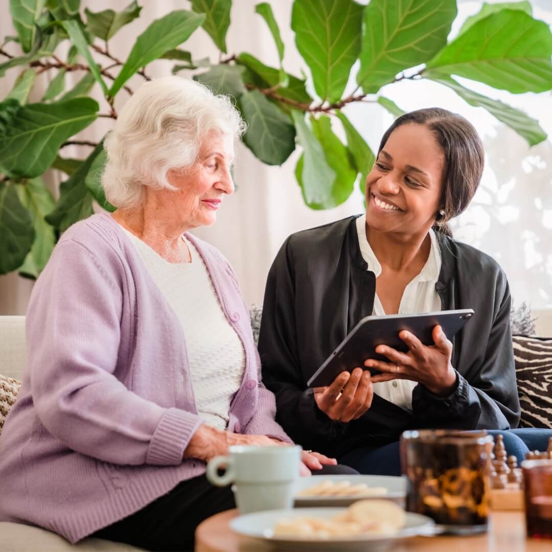 An elderly woman and a younger woman smiling and looking at a tablet together in a cozy living room. - Home Instead