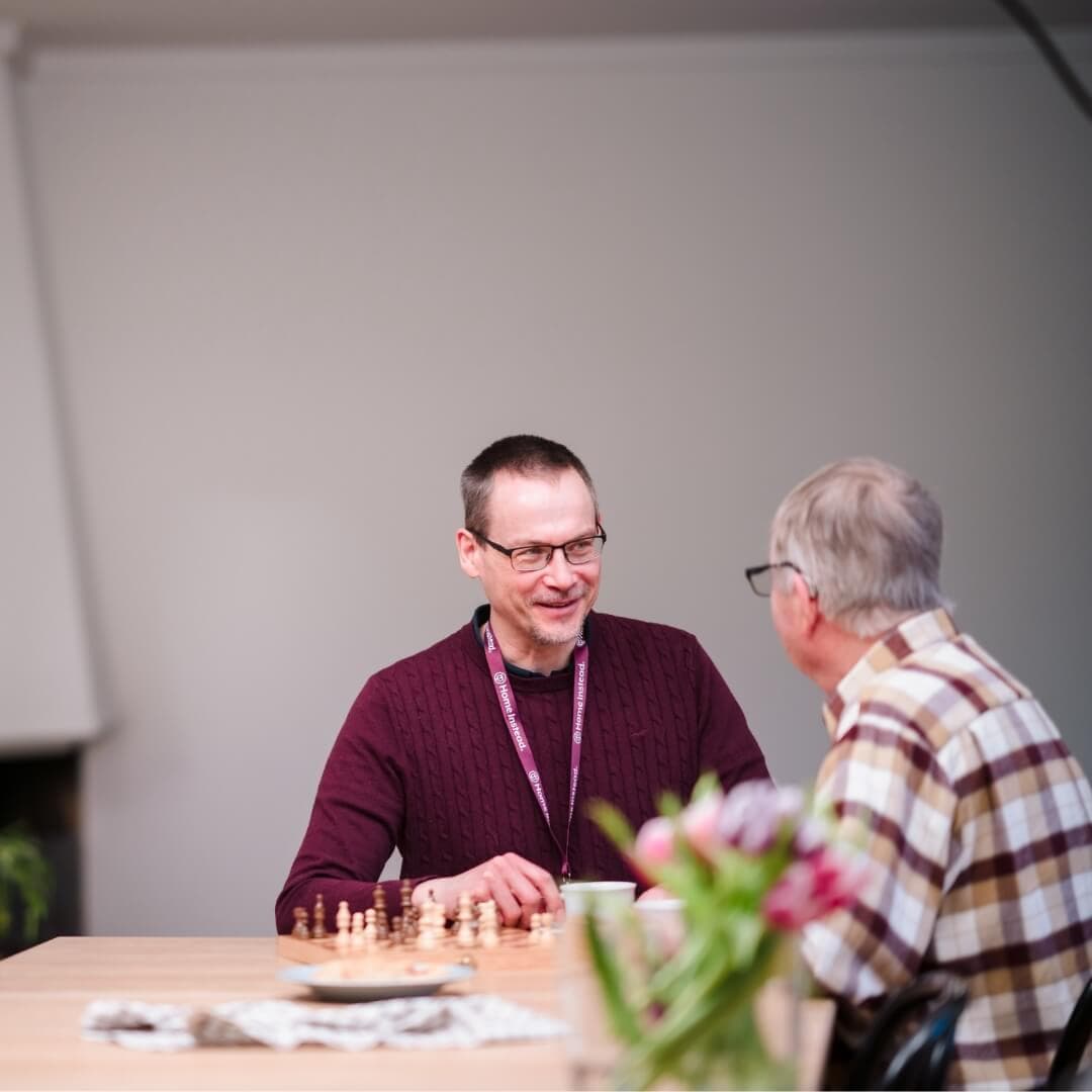 Two men are sitting at a table playing chess and chatting. Tulips are in the foreground on the right side. - Home Instead