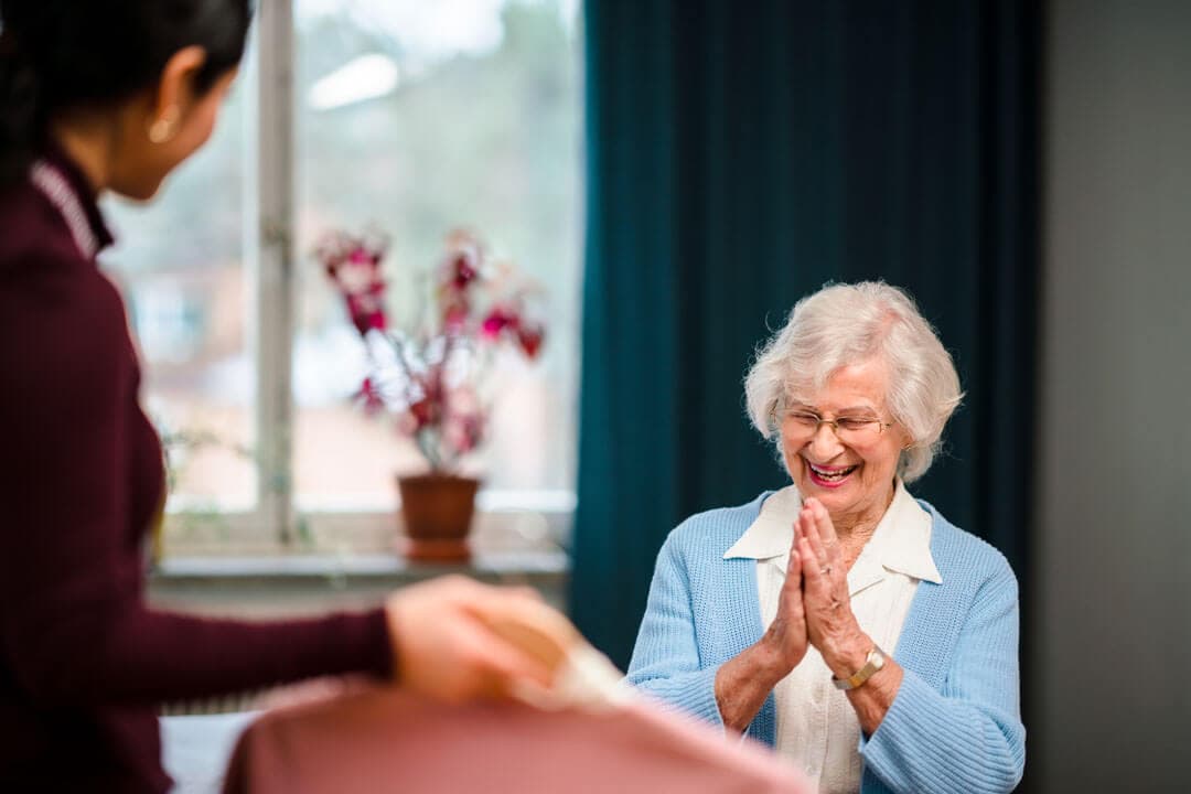 Elderly woman smiling with clasped hands, sitting indoors by window, as another person stands nearby holding a tray. - Home Instead