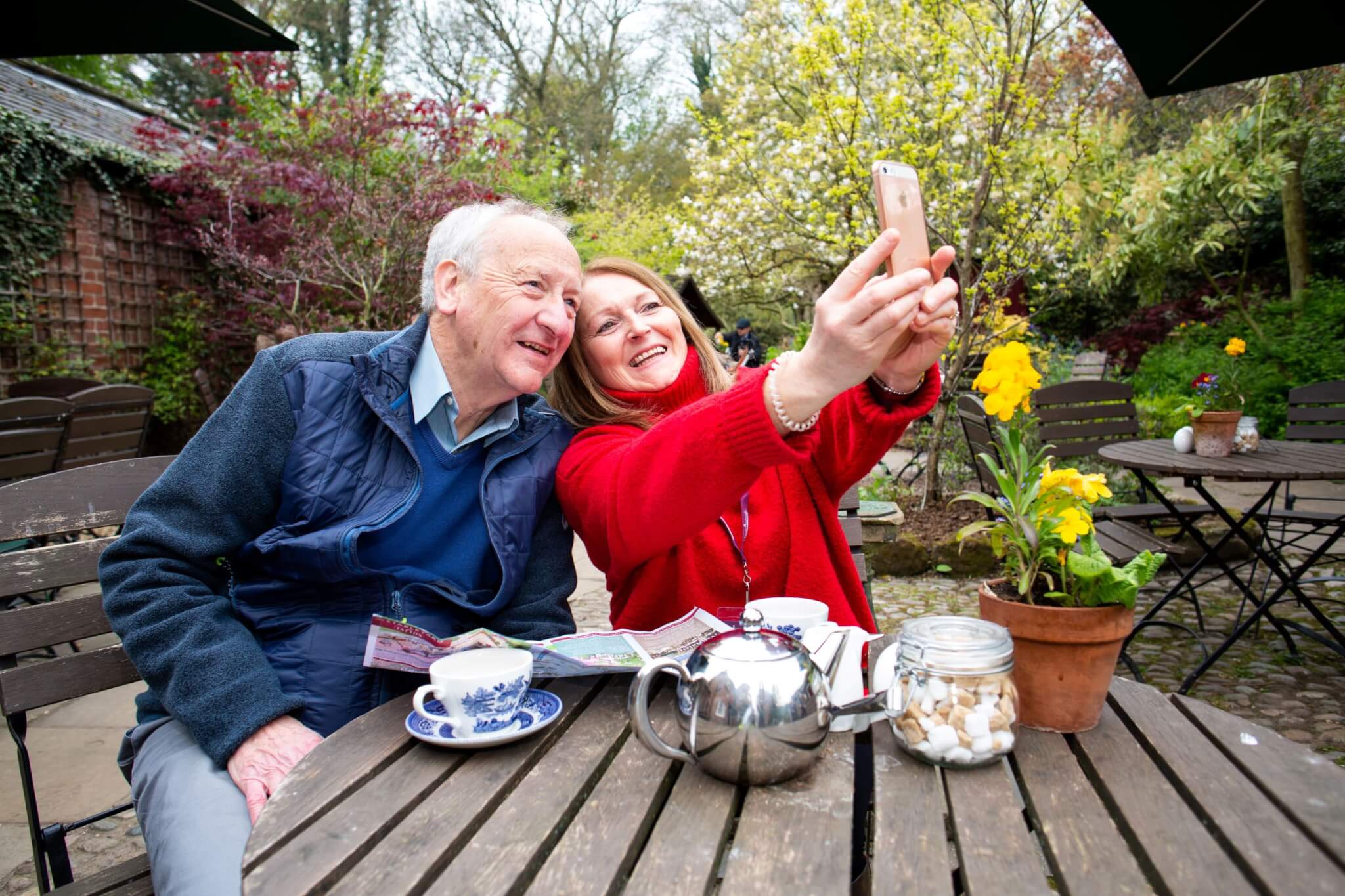 Older man and woman sitting outside at a table, smiling and taking a selfie, with flowers and teacups on the table. - Home Instead