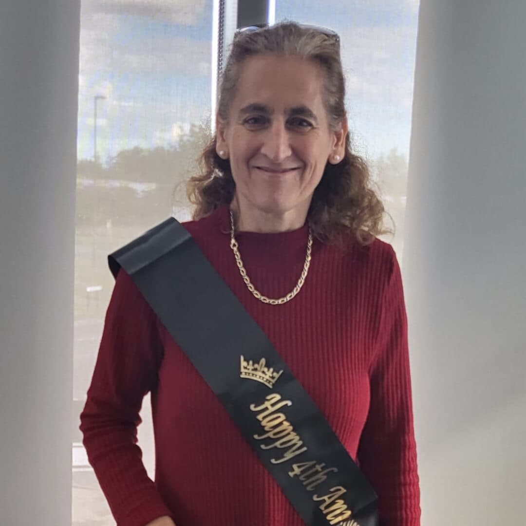 A woman with long brown hair wearing a red sweater and a sash that reads "Happy 4th Anniv" stands, smiling. - Home Instead