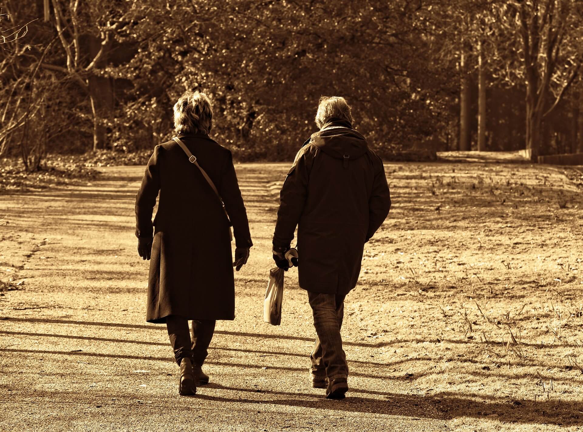 Two people in coats walking on a path in a park with trees, in sepia tone. - Home Instead