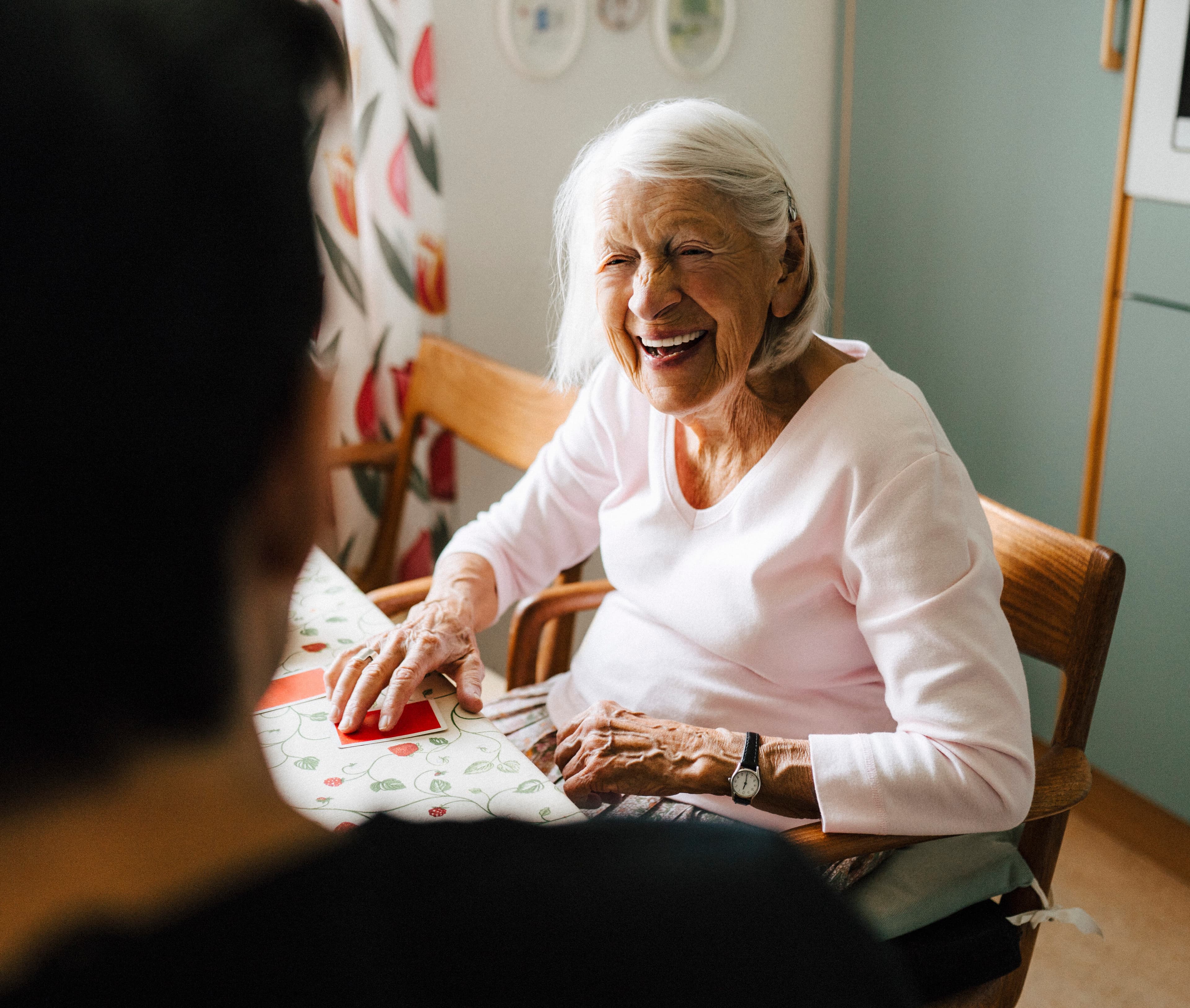 Cheerful senior woman talking with male caregiver while playing cards in kitchen