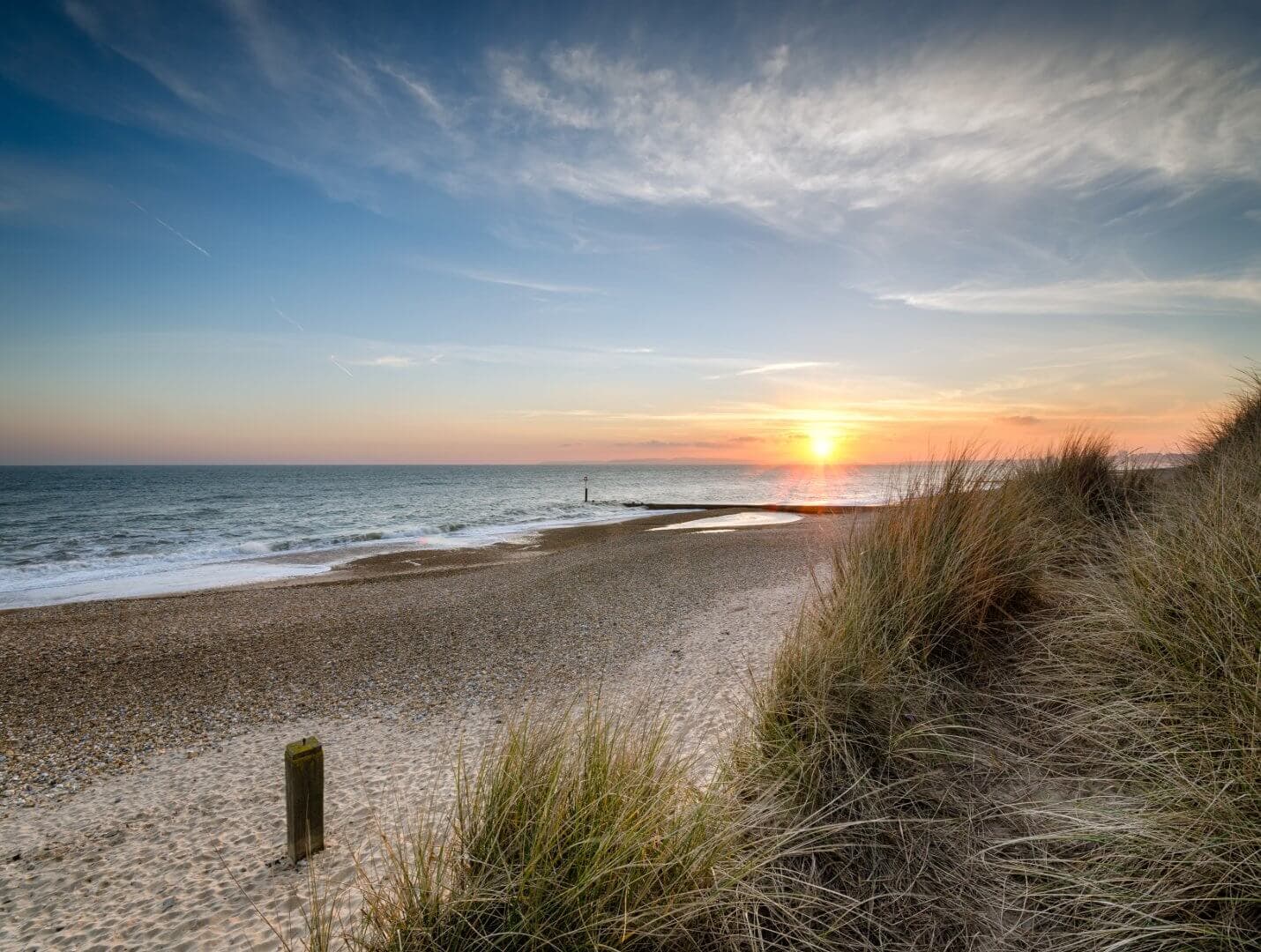 Sunset over a sandy beach with tall grass in the foreground and calm ocean waves under a partly cloudy sky. - Home Instead Bournemouth & Christchurch