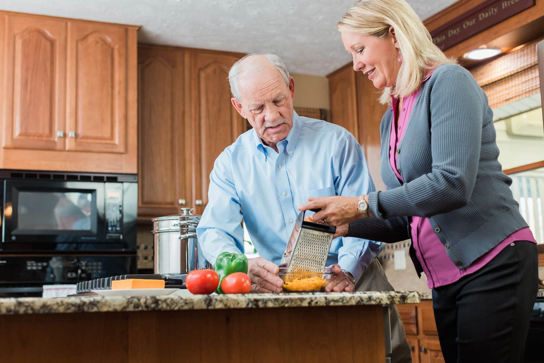 An older couple happily preparing food together in a kitchen, grating cheese on a counter with vegetables nearby. - Home Instead
