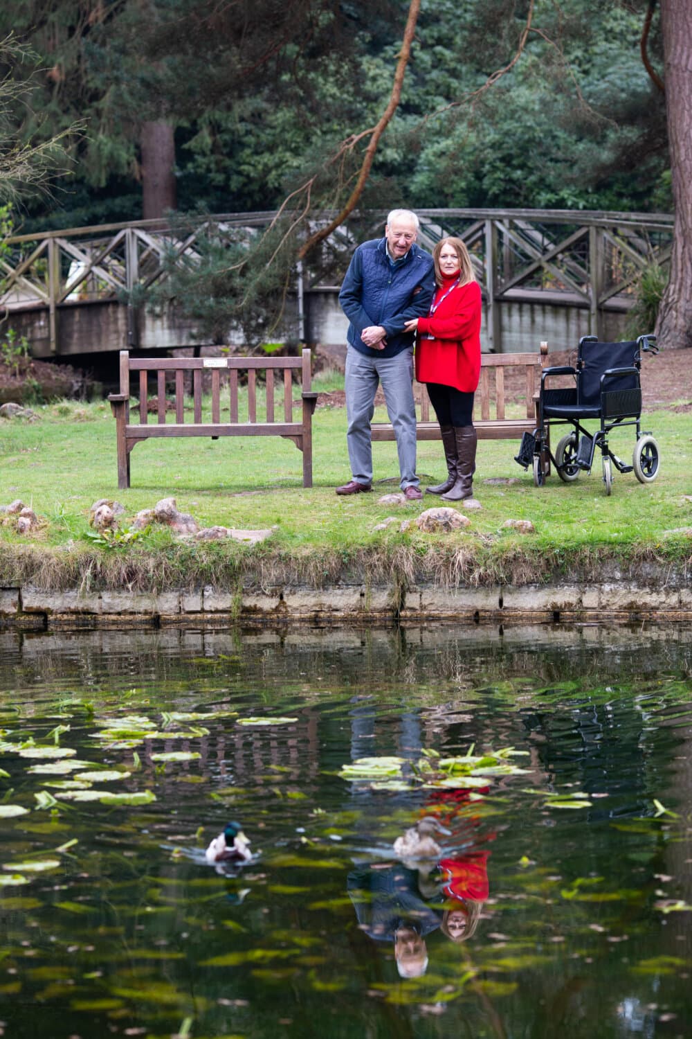 An elderly couple stands by a pond, holding hands. A wheelchair and bench are nearby, with ducks in the pond. - Home Instead