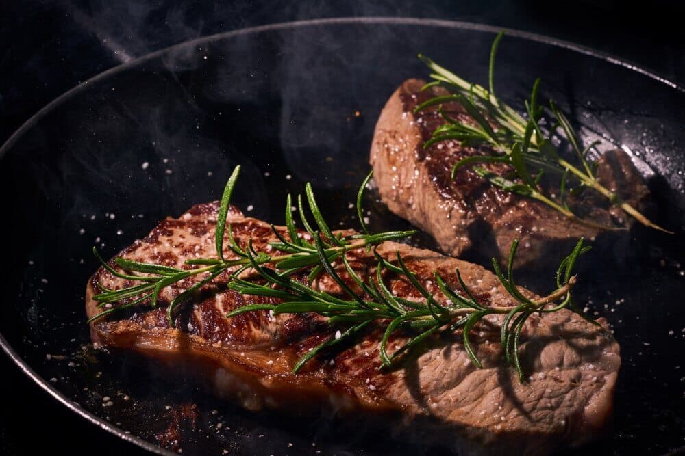Two sizzling steaks in a frying pan, garnished with sprigs of fresh rosemary amidst rising steam. - Home Instead