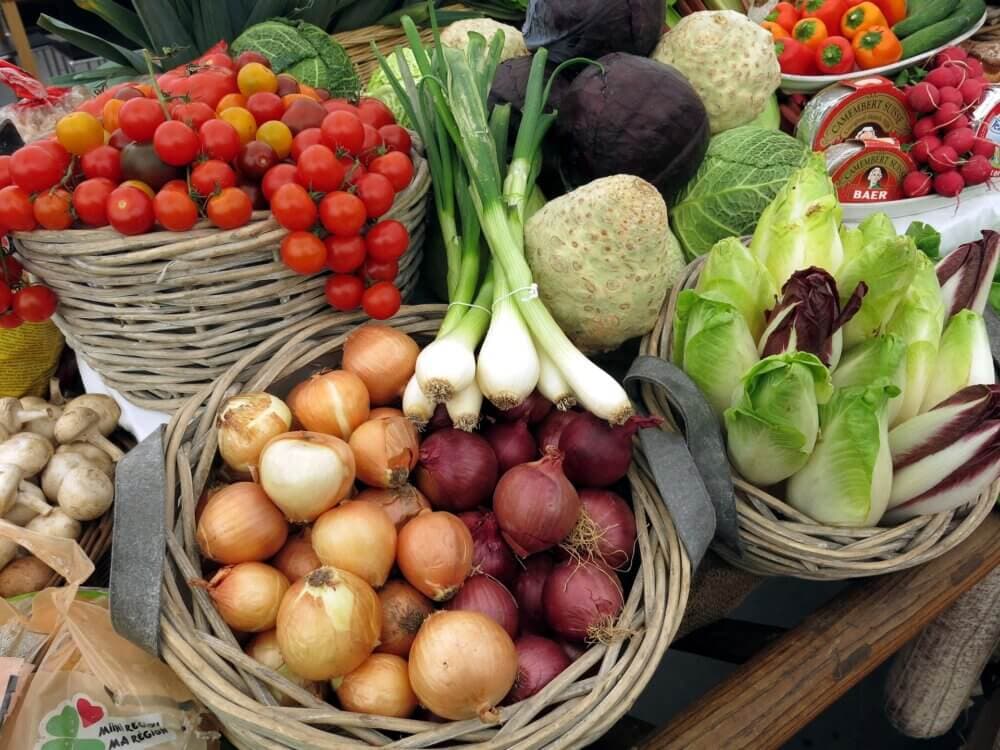 Various fresh vegetables, including tomatoes, onions, leeks, and radicchio, displayed in baskets at a market stall. - Home Instead