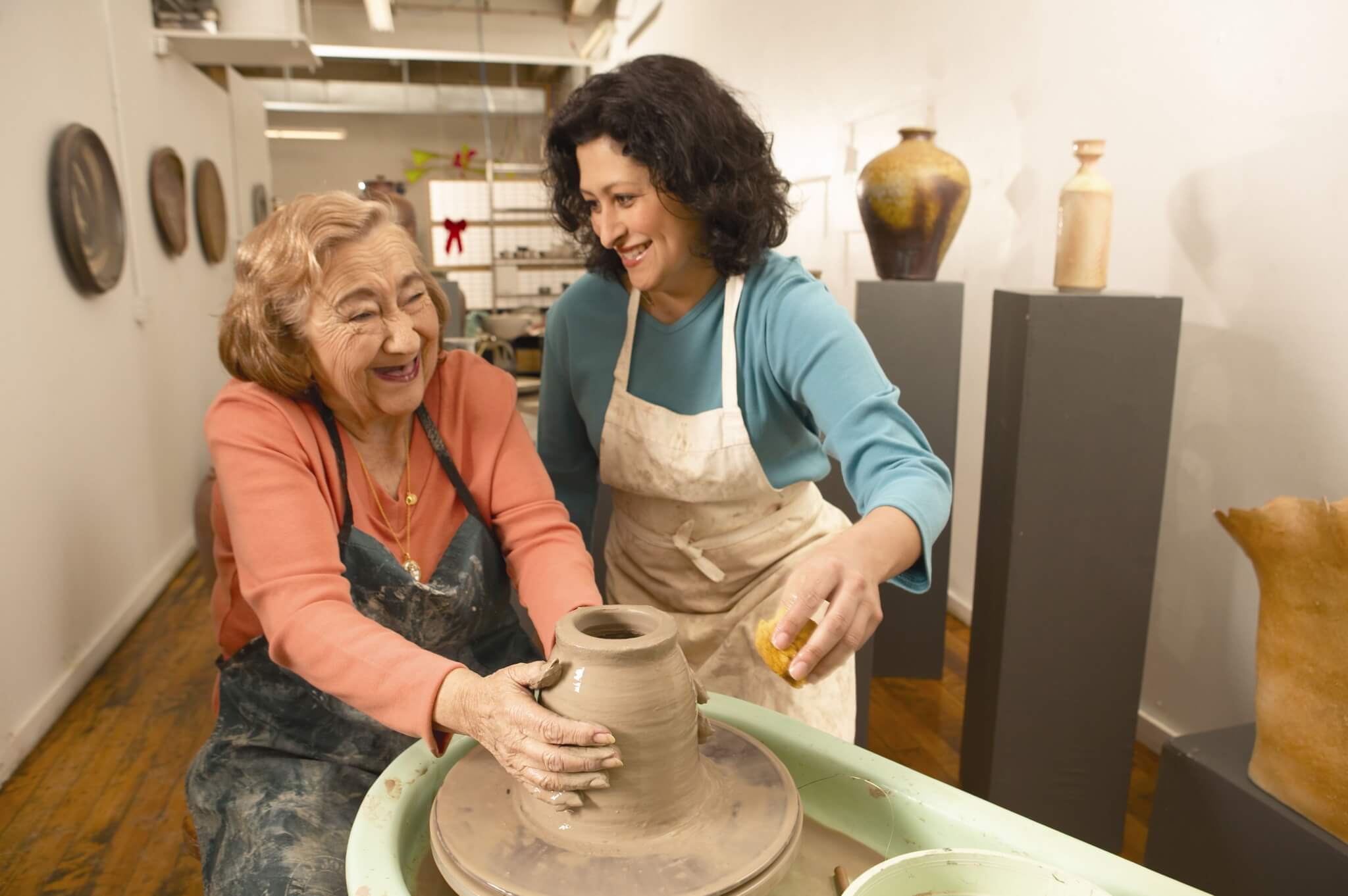 Two women smiling while crafting pottery together in a bright studio. One shapes clay on a pottery wheel, the other assists. - Home Instead