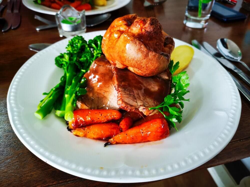 A plate of roast beef with Yorkshire pudding, roasted carrots, and broccoli on a wooden table. - Home Instead