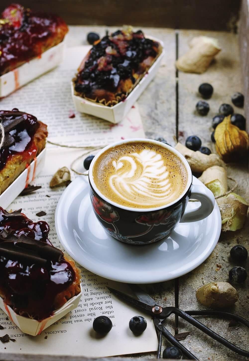 A cup of coffee with latte art in a floral cup, surrounded by pastries, blueberries, and ginger on an open book. - Home Instead