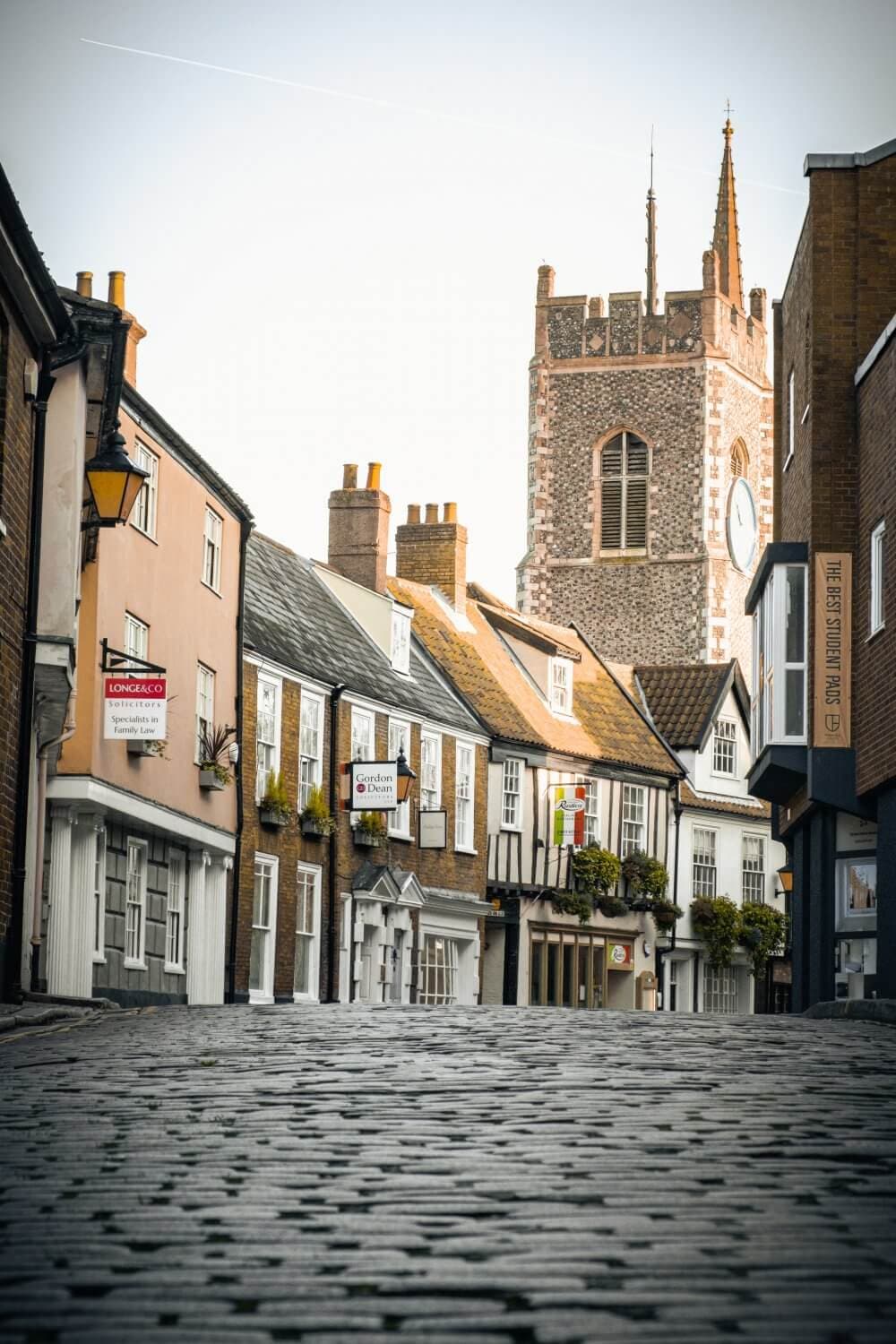 Cobblestone street in a historic town, with old buildings and a tall church tower in the background. - Home Instead