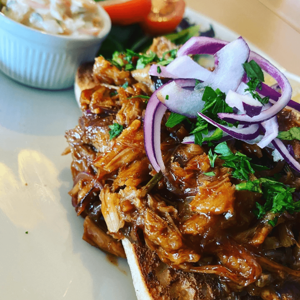 Pulled pork sandwich topped with red onion and herbs, with a side dish of coleslaw and fresh vegetables on the plate. - Home Instead