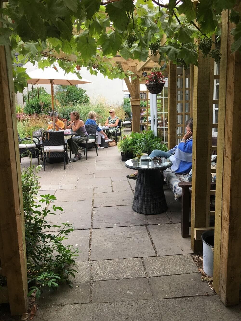 People dining at outdoor tables in a lush garden setting, with greenery overhead and potted plants around. - Home Instead