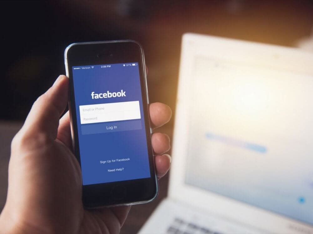 Hand holding a smartphone with the Facebook login screen displayed; a laptop is blurred in the background. - Home Instead