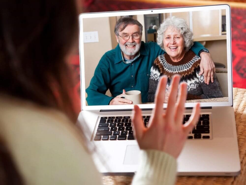 A person waves at an elderly couple smiling on a video call displayed on a laptop screen. - Home Instead