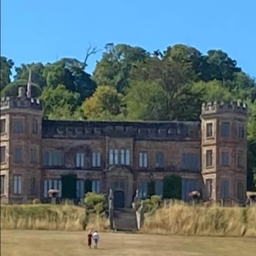 A historic brick mansion with towers, lush greenery in the background, and two people on the lawn in front. - Home Instead