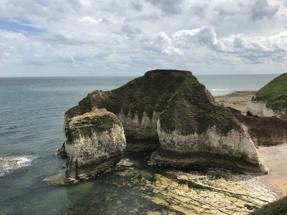 Rocky cliffs covered in greenery jutting into the ocean under a cloudy sky, with a small beach in the background. - Home Instead
