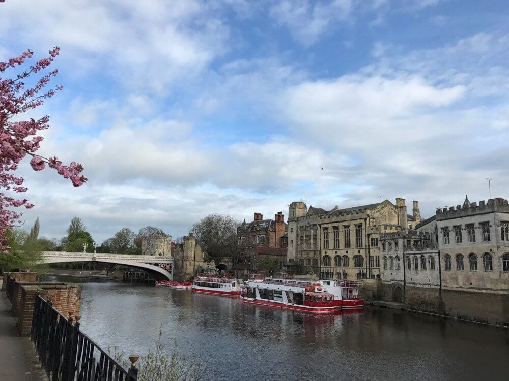 River with boats docked by historic buildings and a bridge; blooming tree on the left under a partly cloudy sky. - Home Instead